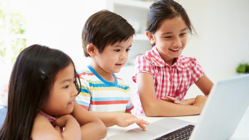 Three children sitting at a table, focused on a laptop screen.