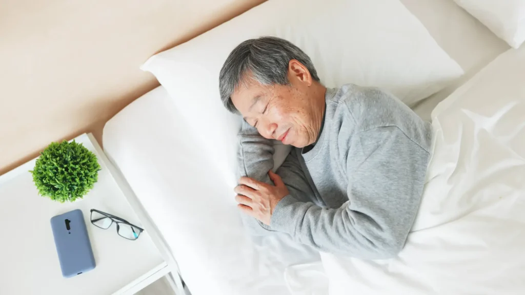 An elderly man peacefully asleep in bed, wearing glasses and holding a phone next to him.