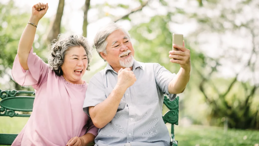 An elderly couple smiling and posing together while taking a selfie with their cell phone.