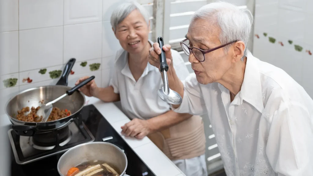 An elderly couple happily cooking together in their cozy kitchen, preparing a delicious meal with love.