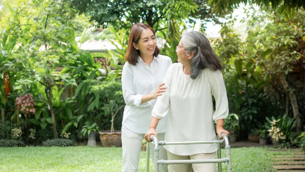 An Asian woman kindly assists an elderly woman with a walker in a beautiful garden.