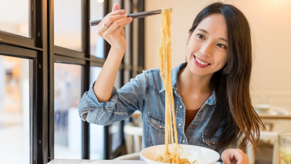 A woman enjoying noodles at a restaurant, savoring every bite with a smile on her face.