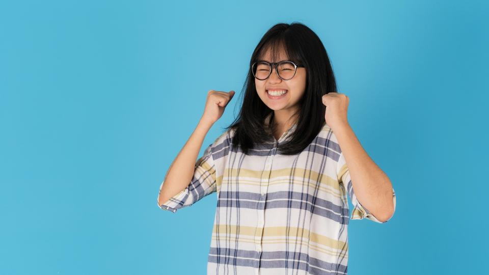 A woman in glasses and a shirt joyfully raises her arms in celebration or victory.