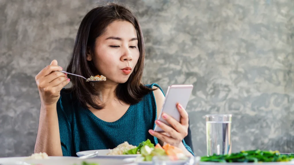 A woman multitasking by eating food and using smartphone.