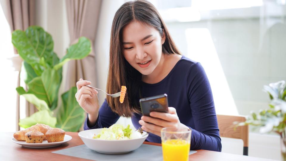 A multitasking woman enjoying a salad while engrossed in her phone, balancing health and technology.