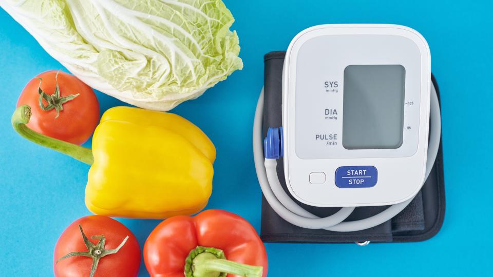 A blood pressure monitor next to a variety of fresh vegetables and a ripe tomato.