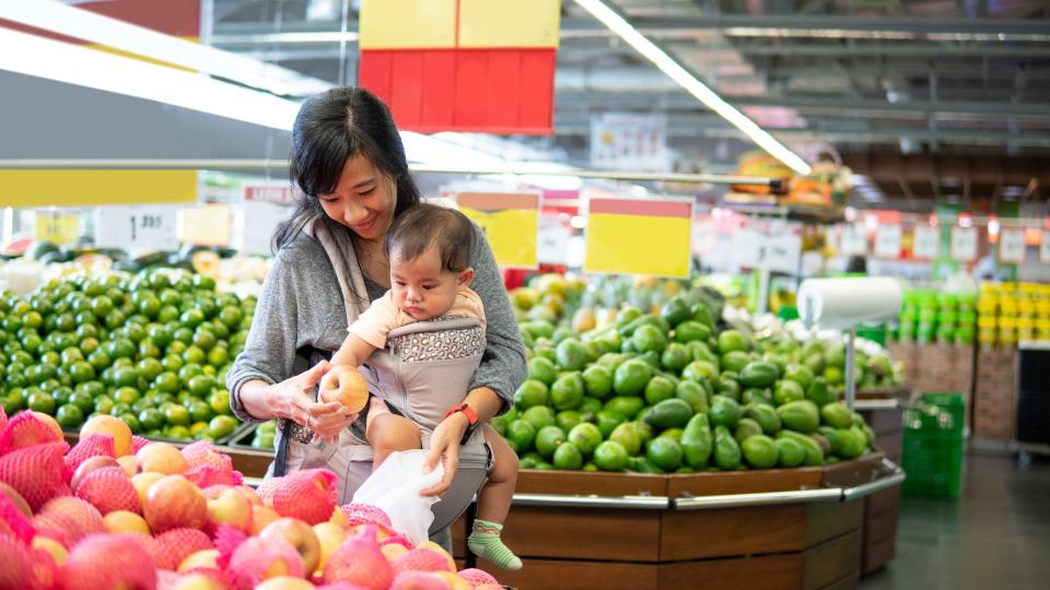 A woman holding a baby in a grocery store, carefully selecting items from the shelves. Postpartum Nutrition