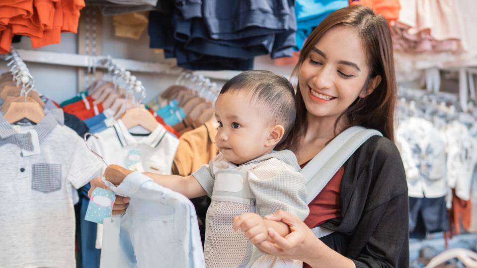 A woman with a baby in a sling gazes at clothes in a store.