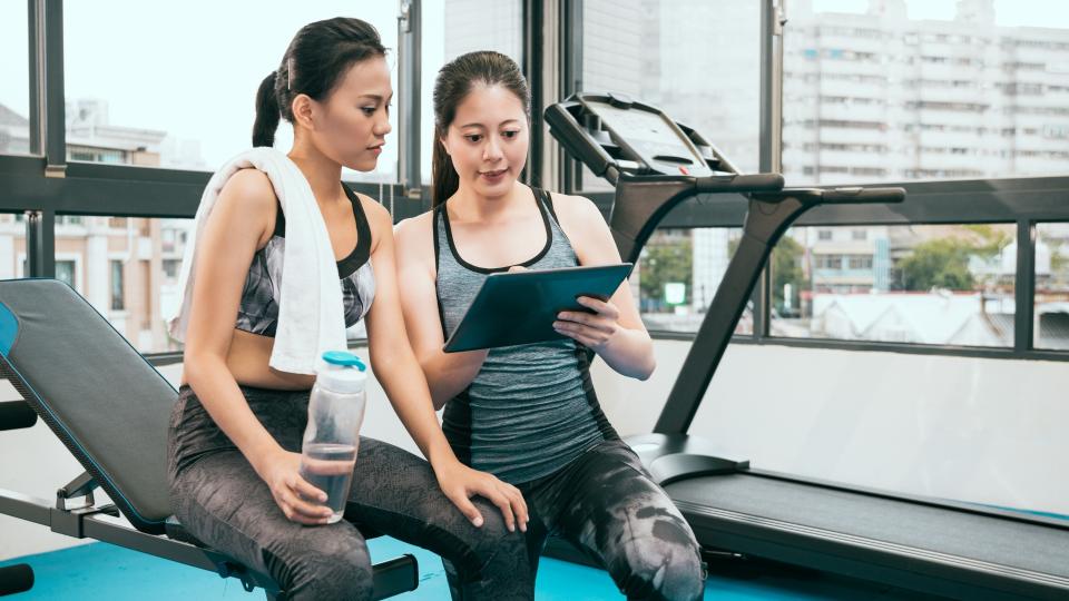 Two women at the gym using a tablet computer to track their workout progress and stay connected. SMART Health Goals