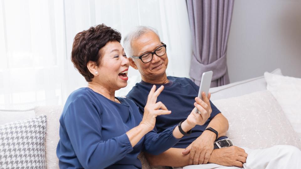 An elderly couple sitting on a couch, looking at a cell phone together.
