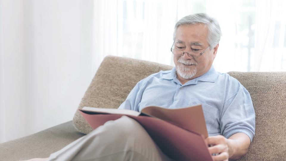 An elderly man sitting on a couch, engrossed in a book.