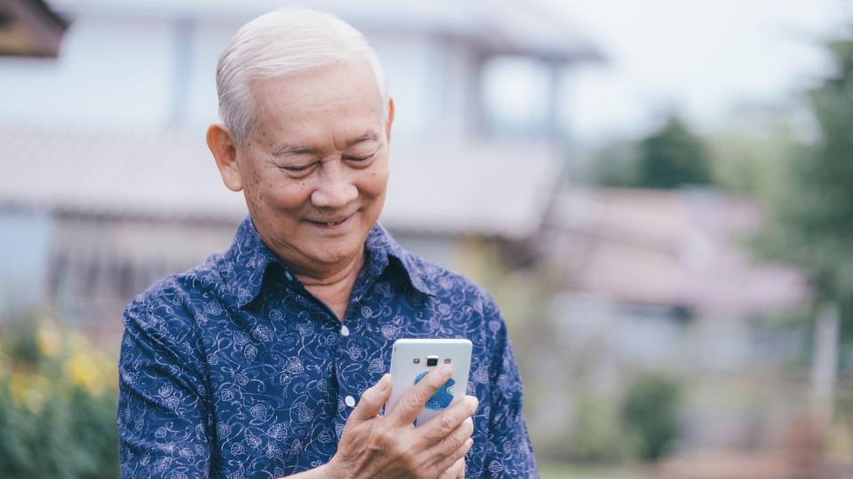 An elderly man sitting on a bench, looking at his cell phone.