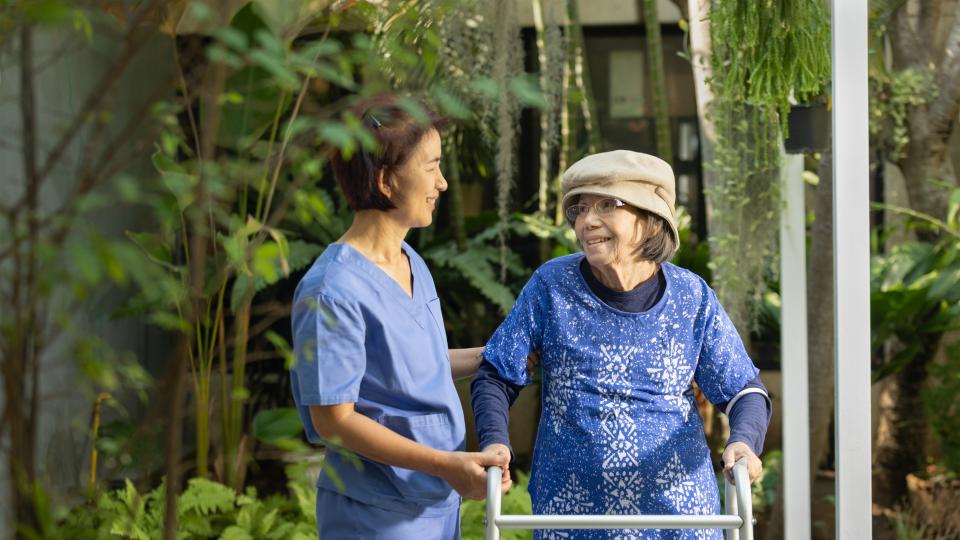 Female healthcare worker in blue scrubs assisting elderly woman with walker.