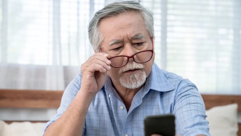 An elderly man wearing glasses intently focuses on his phone screen, engrossed in whatever he is viewing or reading. eyesight problems