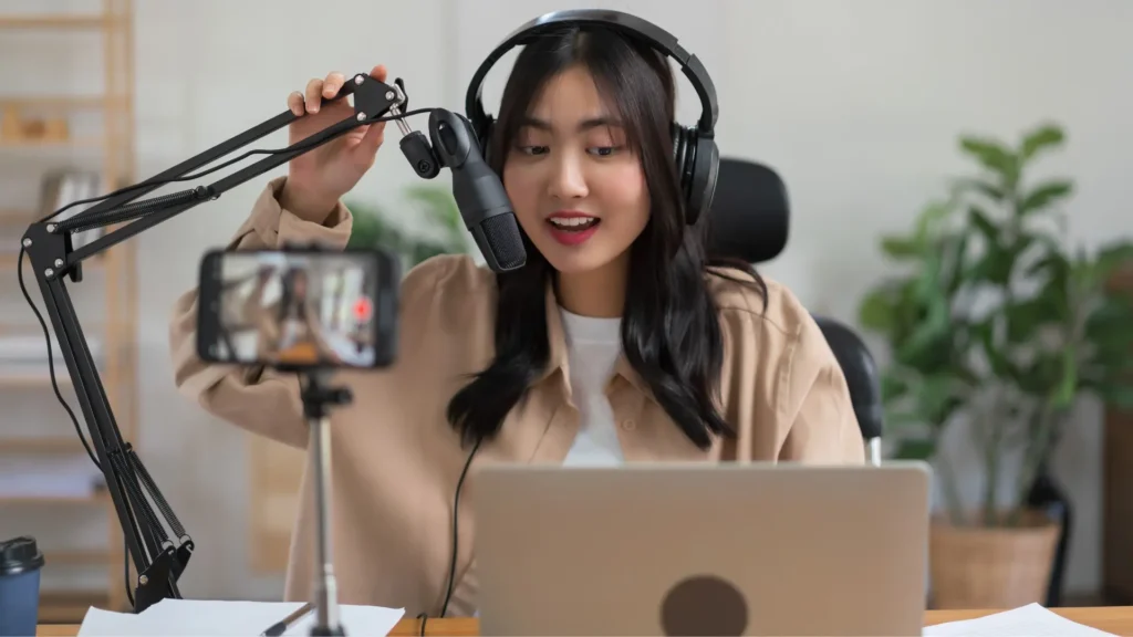 Woman in headphones holding up microphone and laptop, ready to record audio for a podcast.