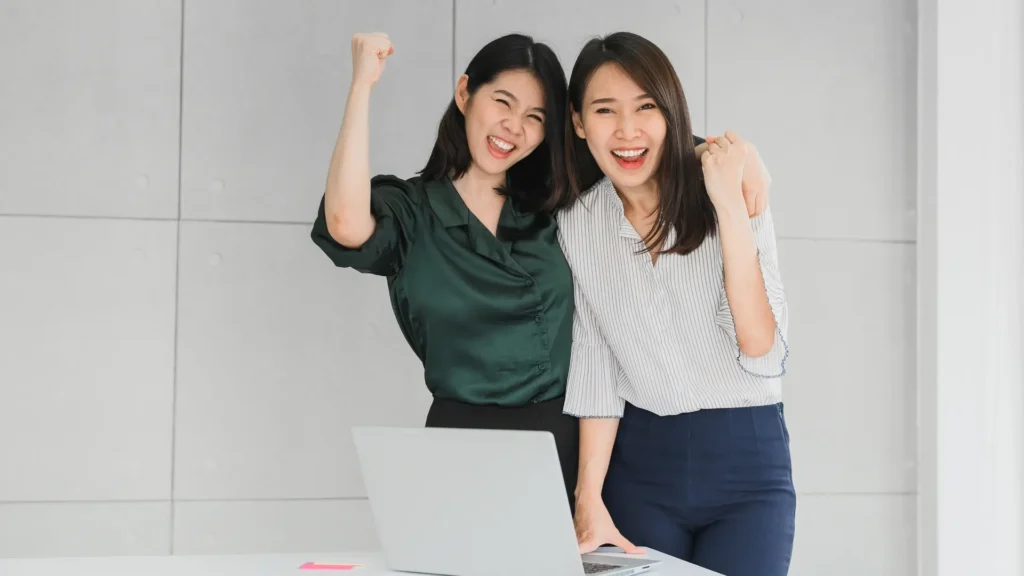 Two women smiling and cheering in front of a laptop screen.