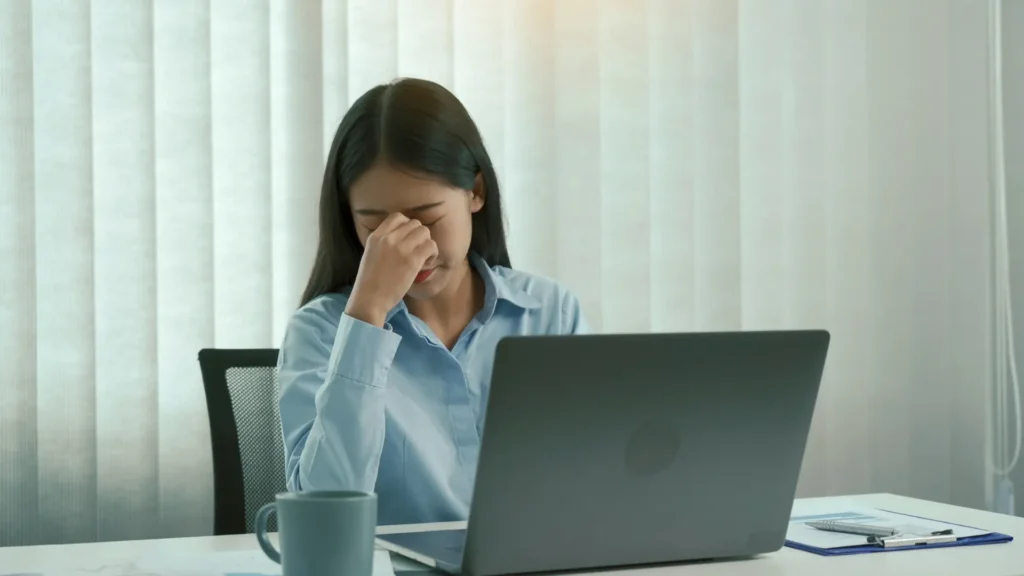 A tired woman sitting at a desk, feeling overwhelmed and stressed, with her head resting in her hands.