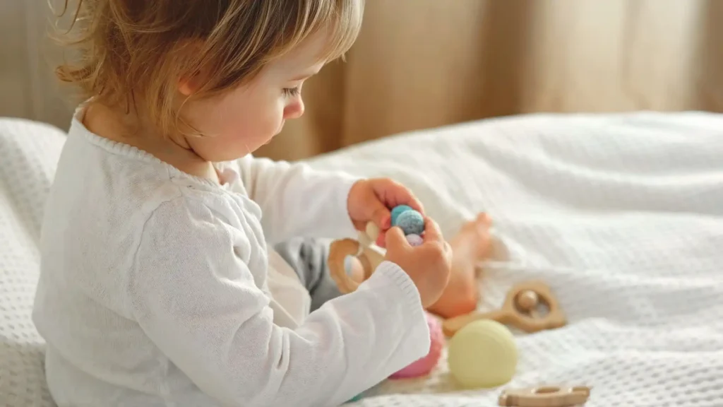 A toddler happily playing with toys on a bed, surrounded by stuffed animals and colorful objects.
