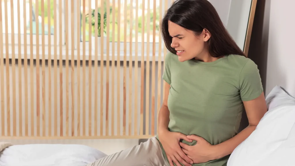 A woman sitting on a bed, clutching her stomach in pain. Postpartum Pain in Pelvic Area