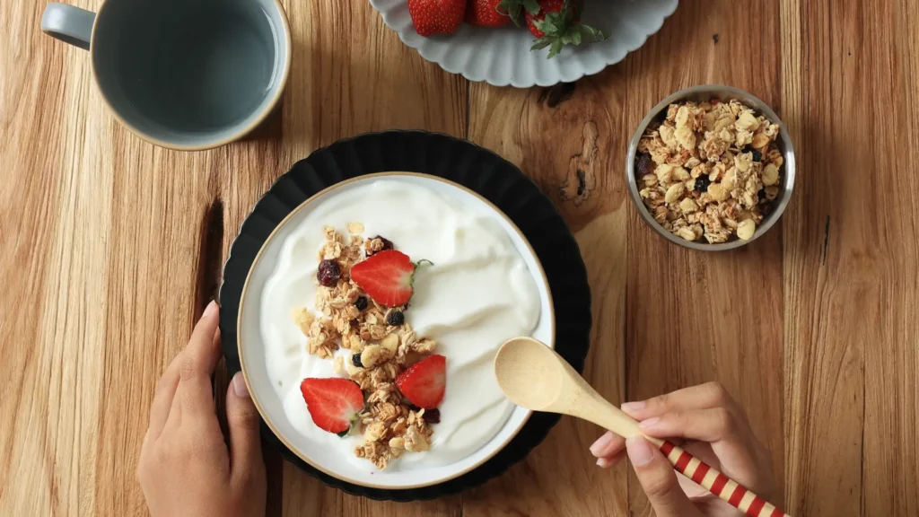 A bowl of yogurt topped with fresh strawberries and granola on a rustic wooden table.