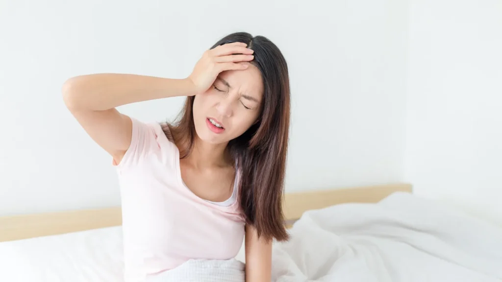 A woman sitting on her bed, holding her head in pain.