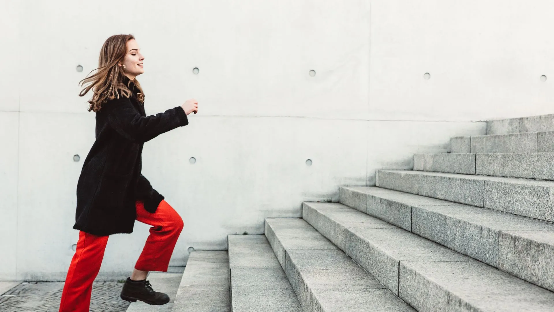 Woman in red pants and black coat walking up wide concrete stairs against a white wall. Climb five flights of stairs daily to lower your heart disease risk by 20%.