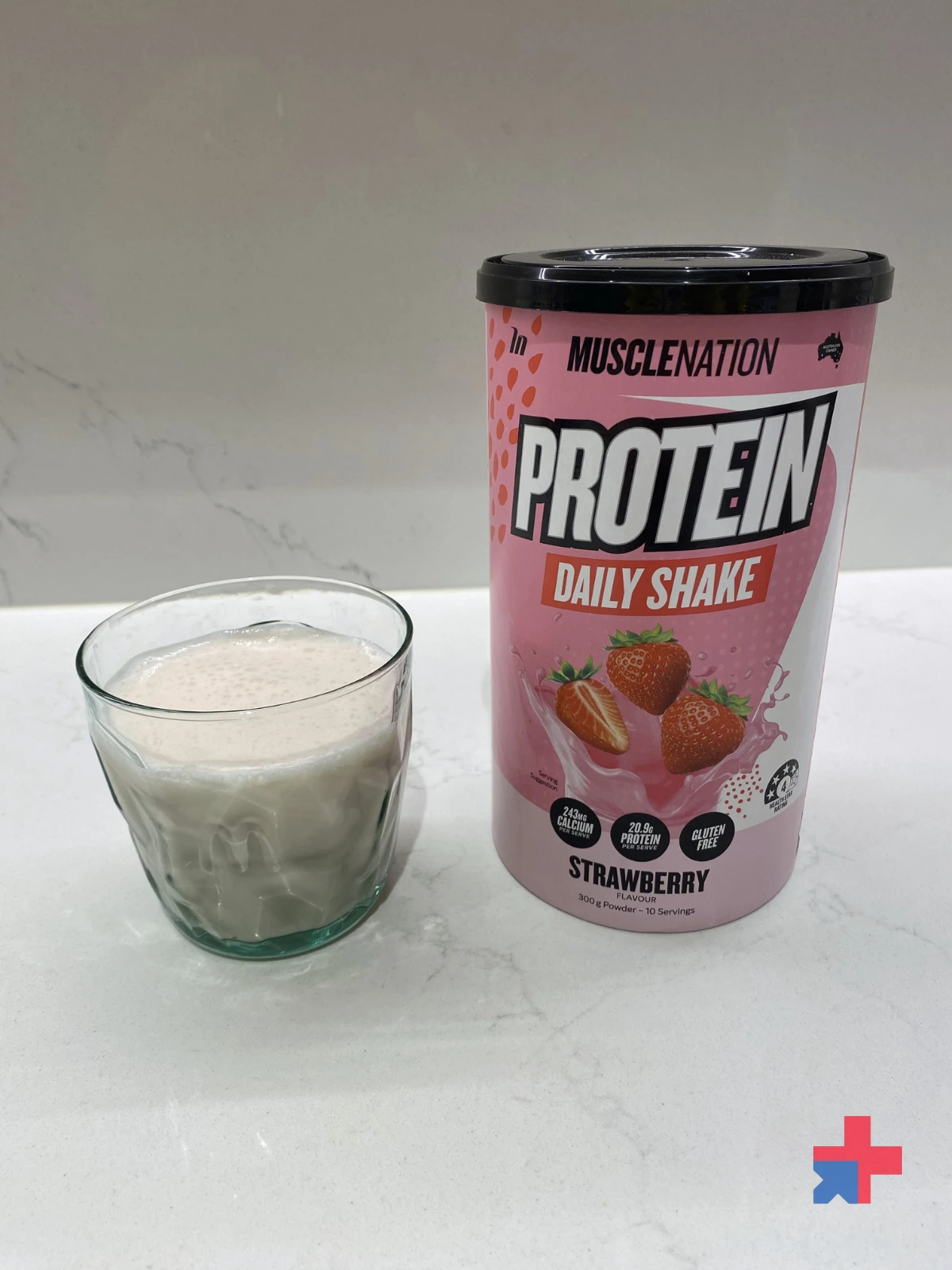 Glass of prepared Muscle Nation Strawberry protein shake beside the pink Protein Daily Shake tub