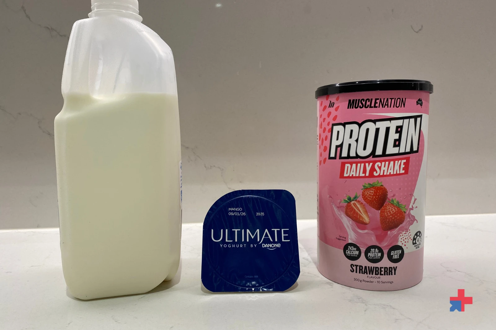 Milk carton, mango yoghurt tub and Muscle Nation Protein Daily Shake Strawberry powder lined up on a kitchen bench