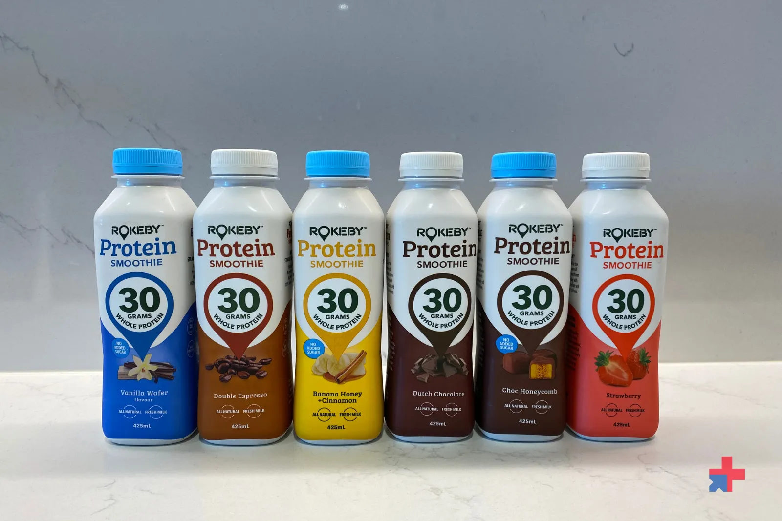 Six Rokeby Protein Smoothie bottles in a row on a white counter, showing Vanilla Wafer, Double Espresso, Banana Honey Cinnamon, Dutch Chocolate, Choc Honeycomb, and Strawberry flavours.