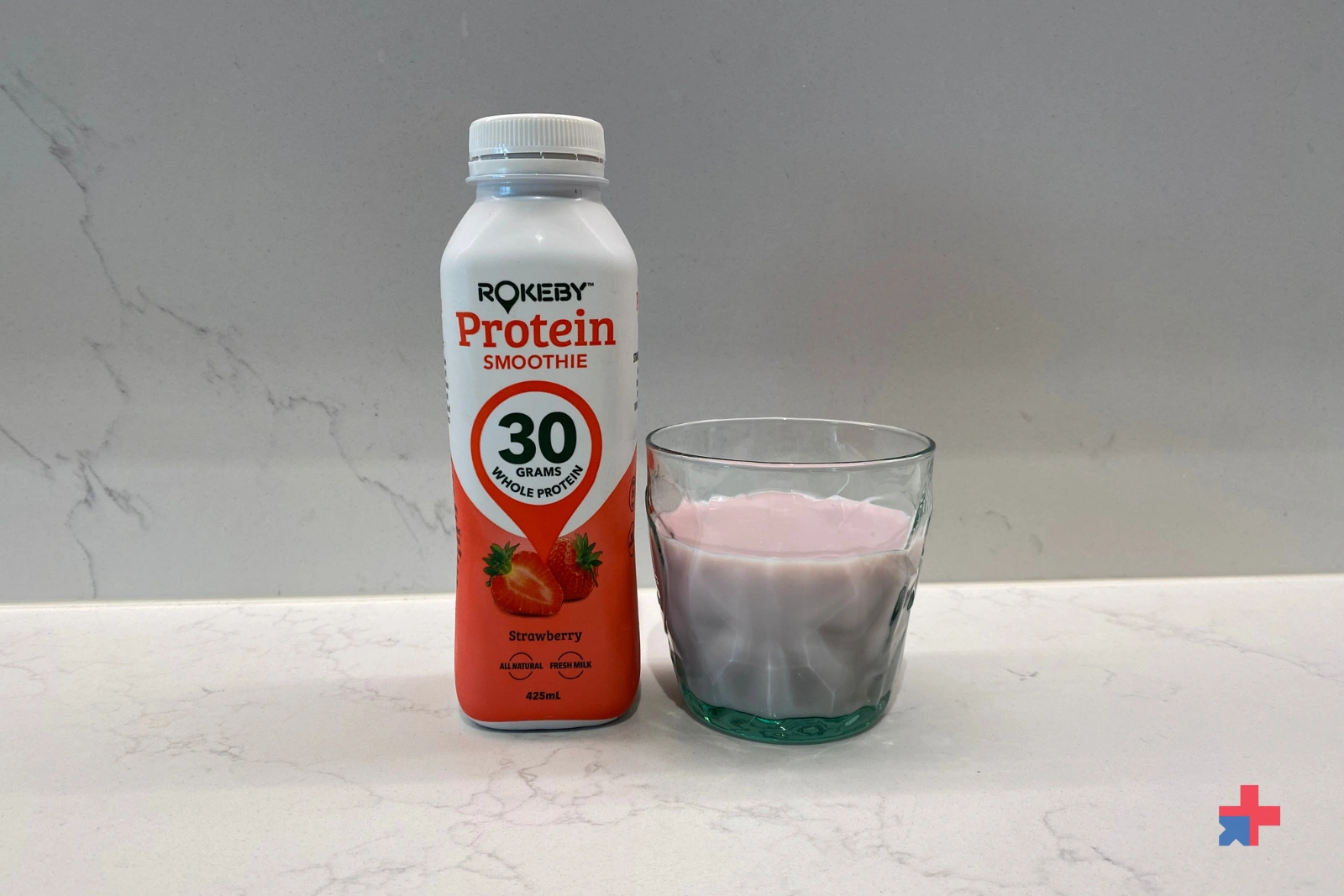 A bottle of Rokeby strawberry protein smoothie in a glass on a white marble countertop.
