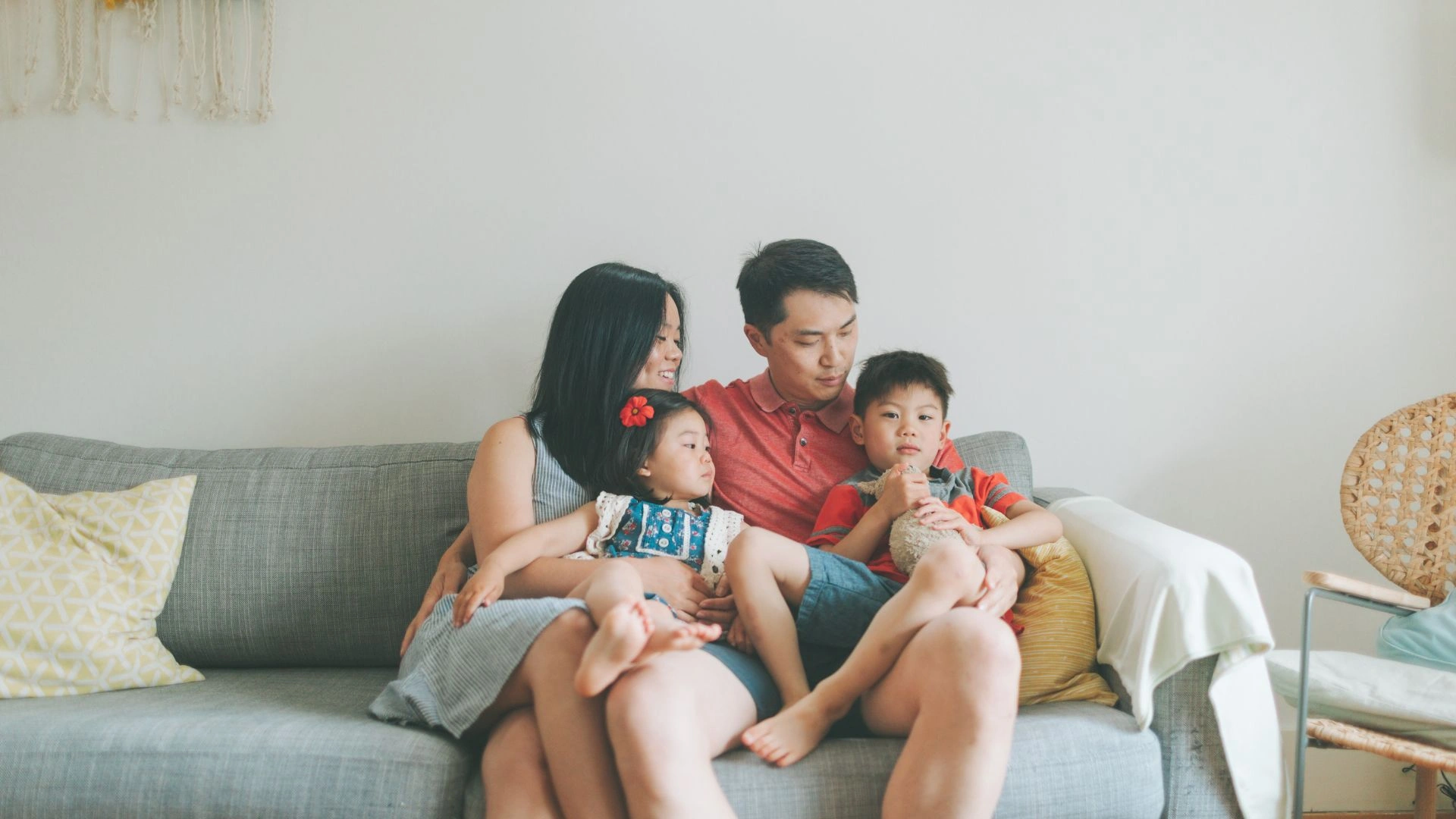 A family of four sits on a gray sofa at home: mother, father, a young girl with a red flower in her hair, and a boy holding a teddy bear.