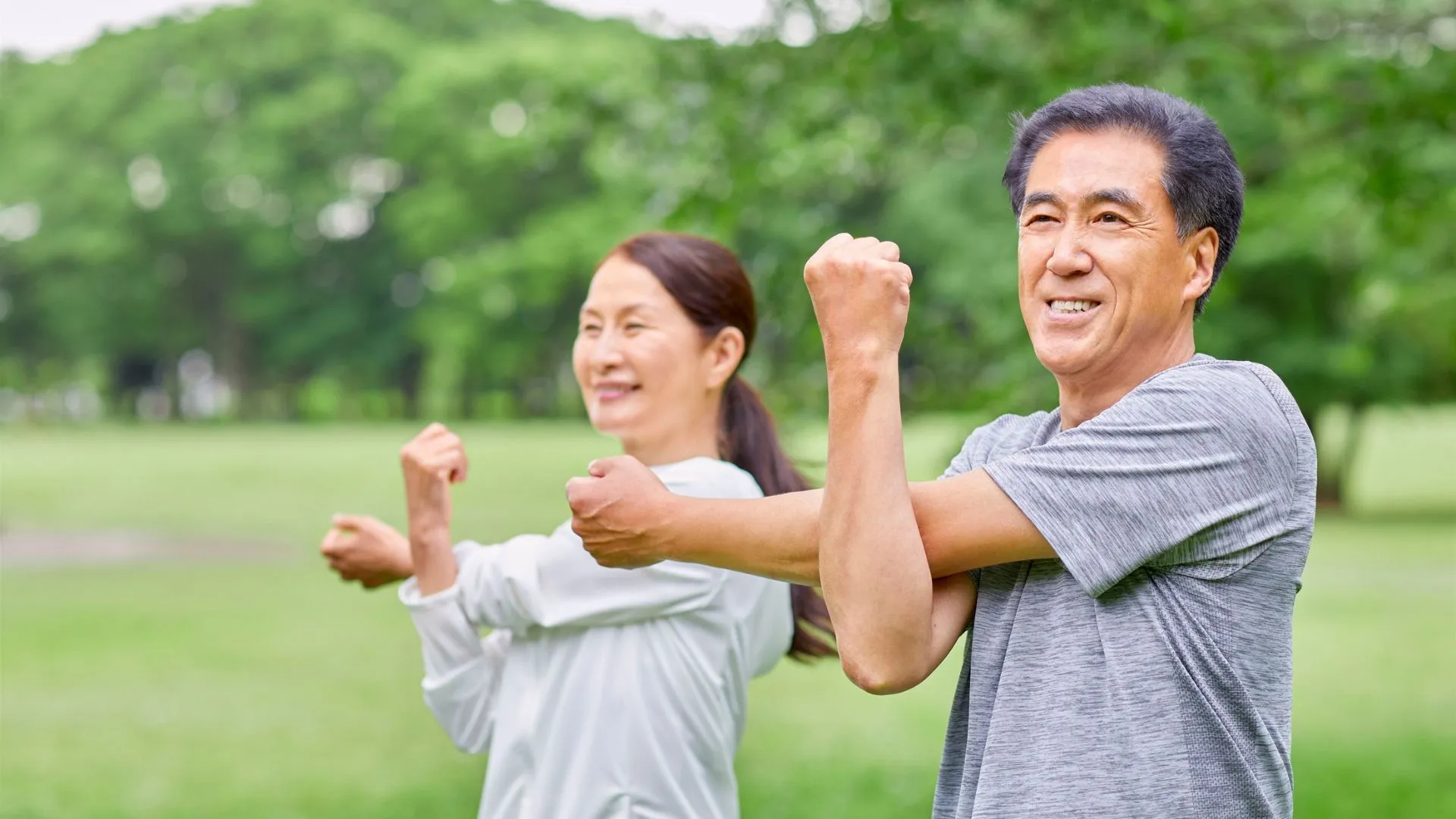Middle-aged Asian couple stretching outdoors in a park, showing light supervised exercise during recovery after a heart attack.