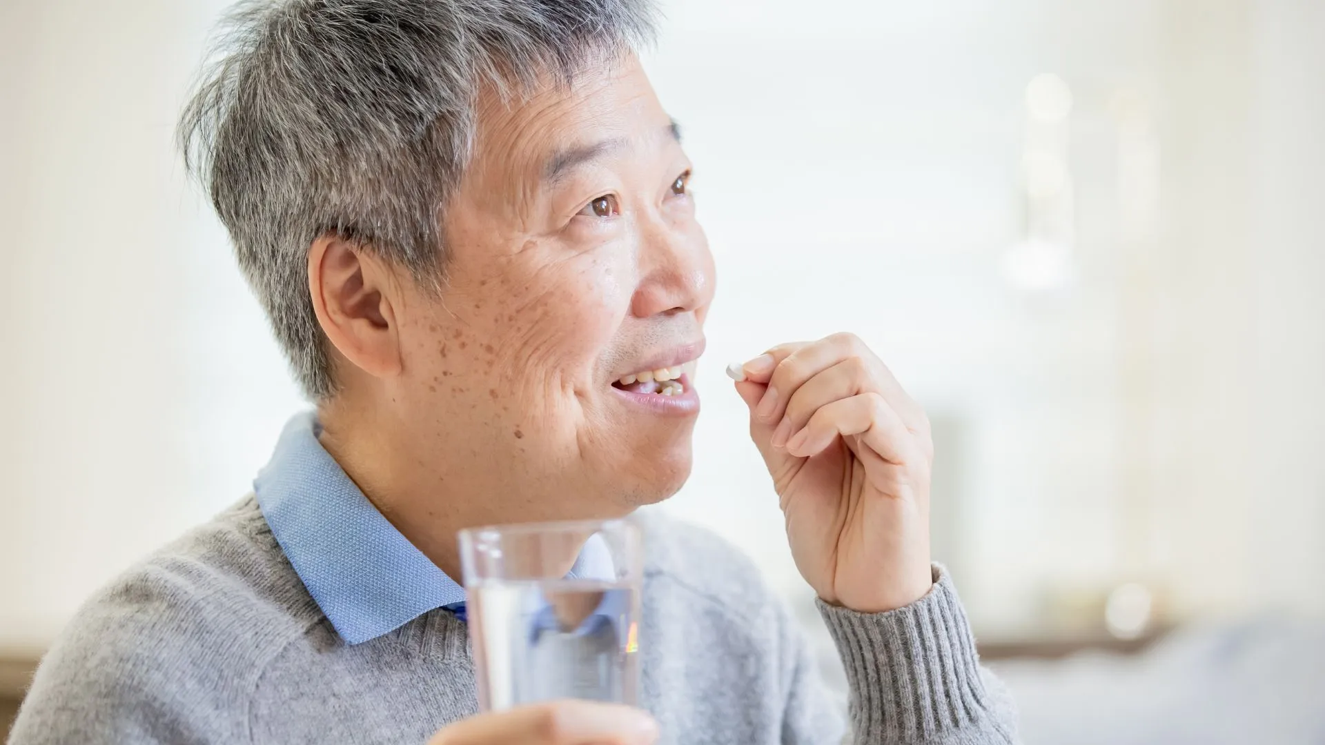 Older Asian man holding a glass of water and taking a small tablet, illustrating daily heart-attack medications such as antiplatelets after stent insertion.