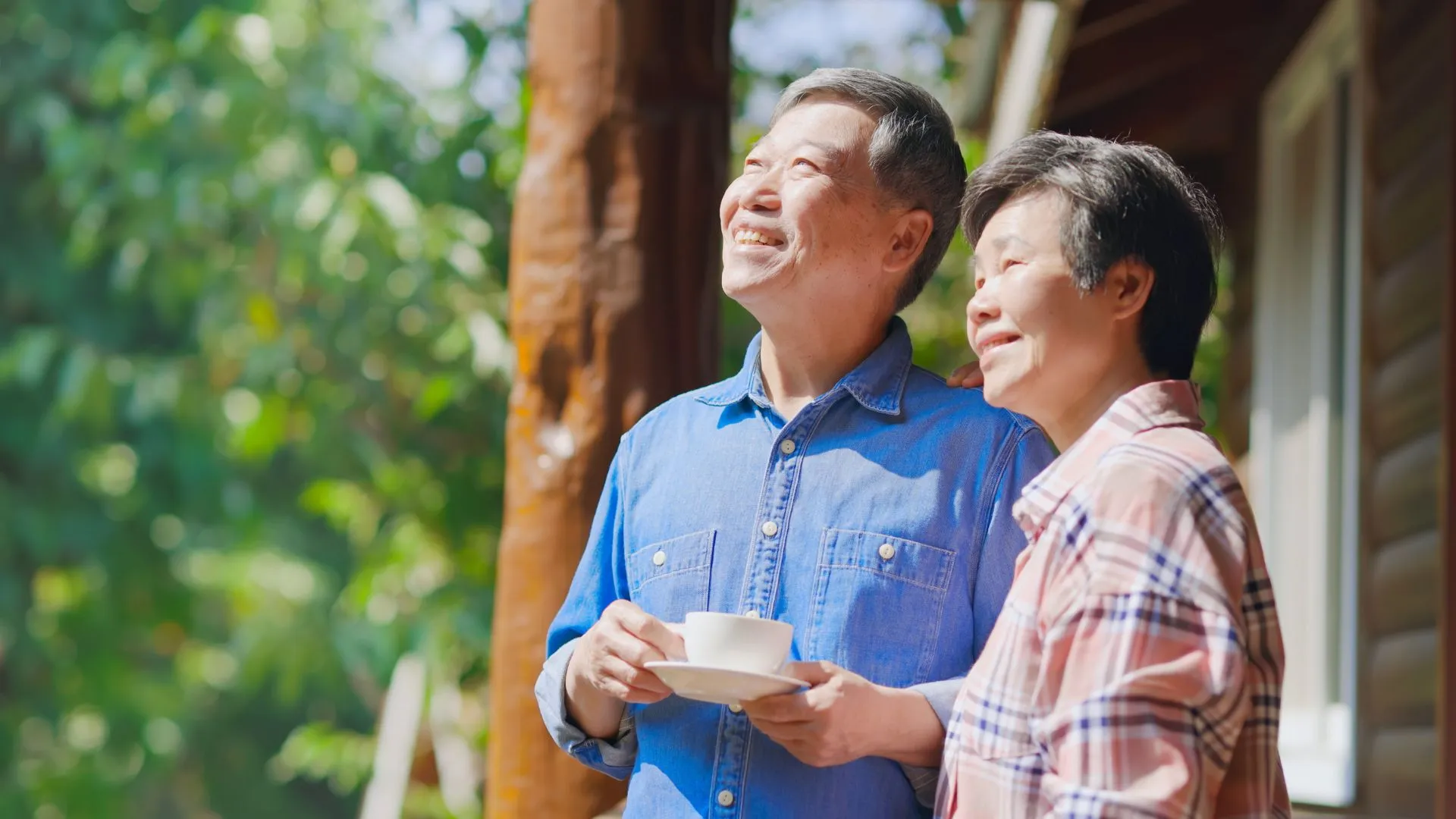 Older Asian couple standing outside a house and smiling, the man holding a cup of coffee, conveying a return to everyday life after a heart attack.