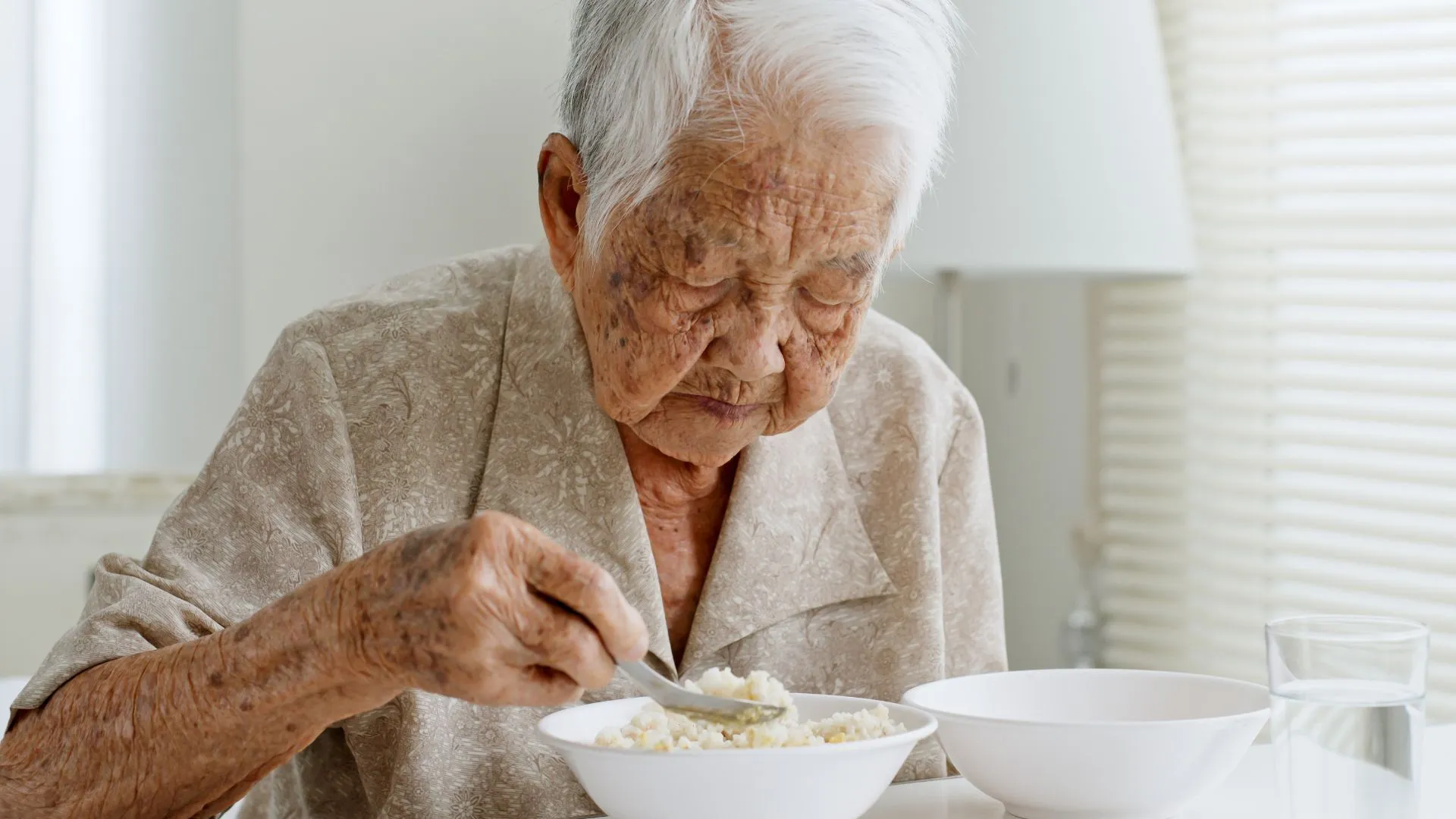 Elderly Asian woman eating a bowl of rice porridge at a table, representing a low-salt congee option often recommended after a heart attack in Singapore.